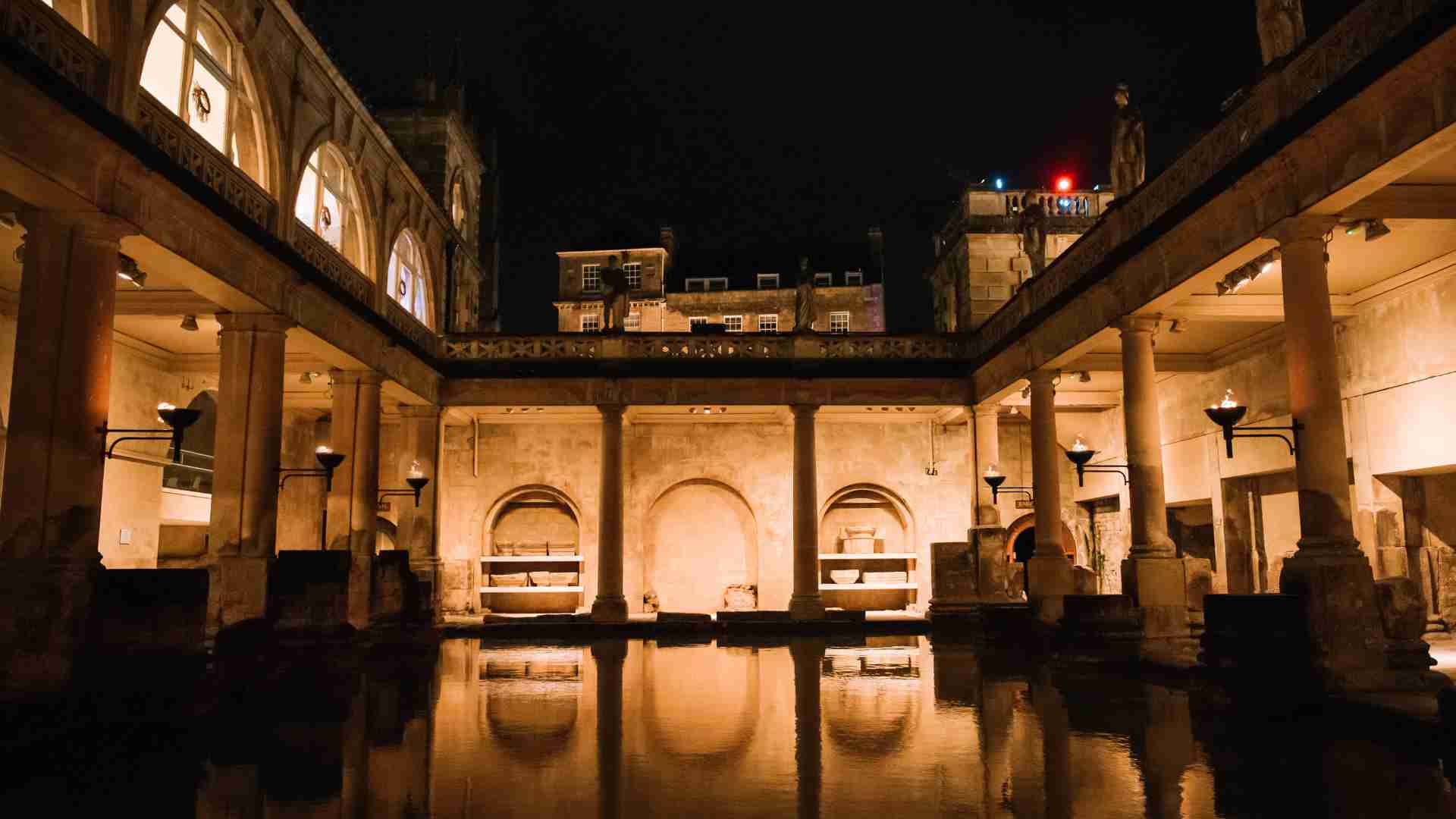 The historic Great Bath at the Roman Baths is illuminated by torches at night, showcasing ancient columns and architecture reflected in the water. This atmospheric setting serves as the location for welcome drinks during the Roman Baths Silent Disco events.