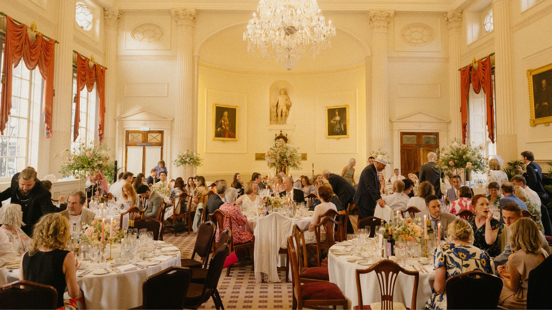 The room features elegant classical architecture with white walls, decorative columns, and a large crystal chandelier hanging from the centre of the ceiling. Numerous guests are seated at round tables covered with white tablecloths.